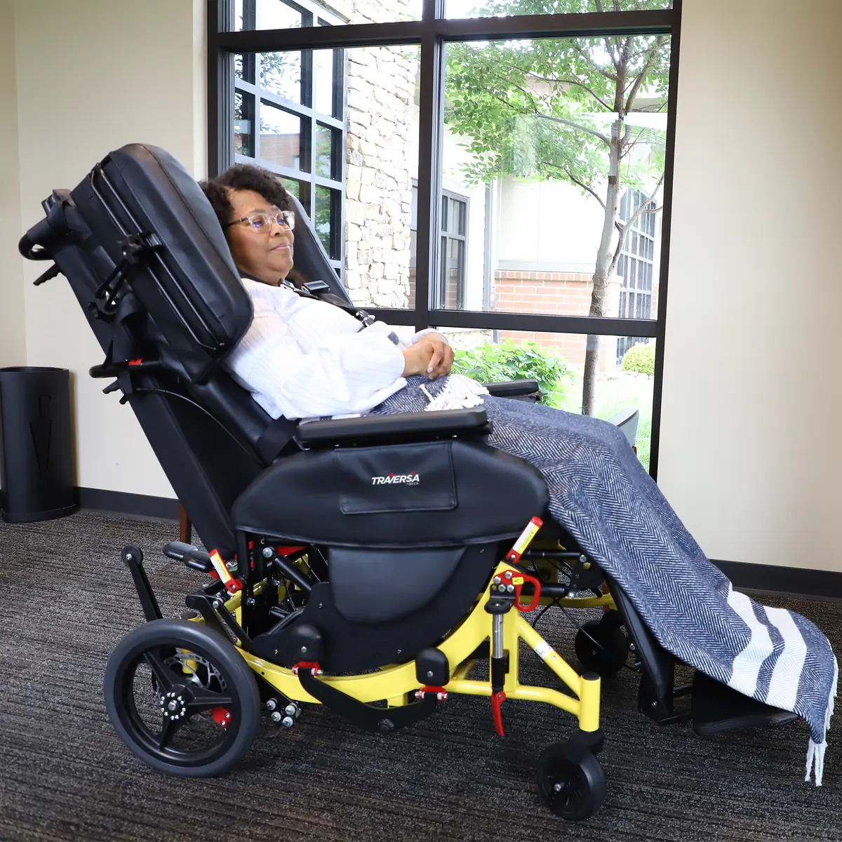 The Broda Traversa reclining transport chair seen from the side, its seat and back tilted together, with a rider resting under a blanket.
