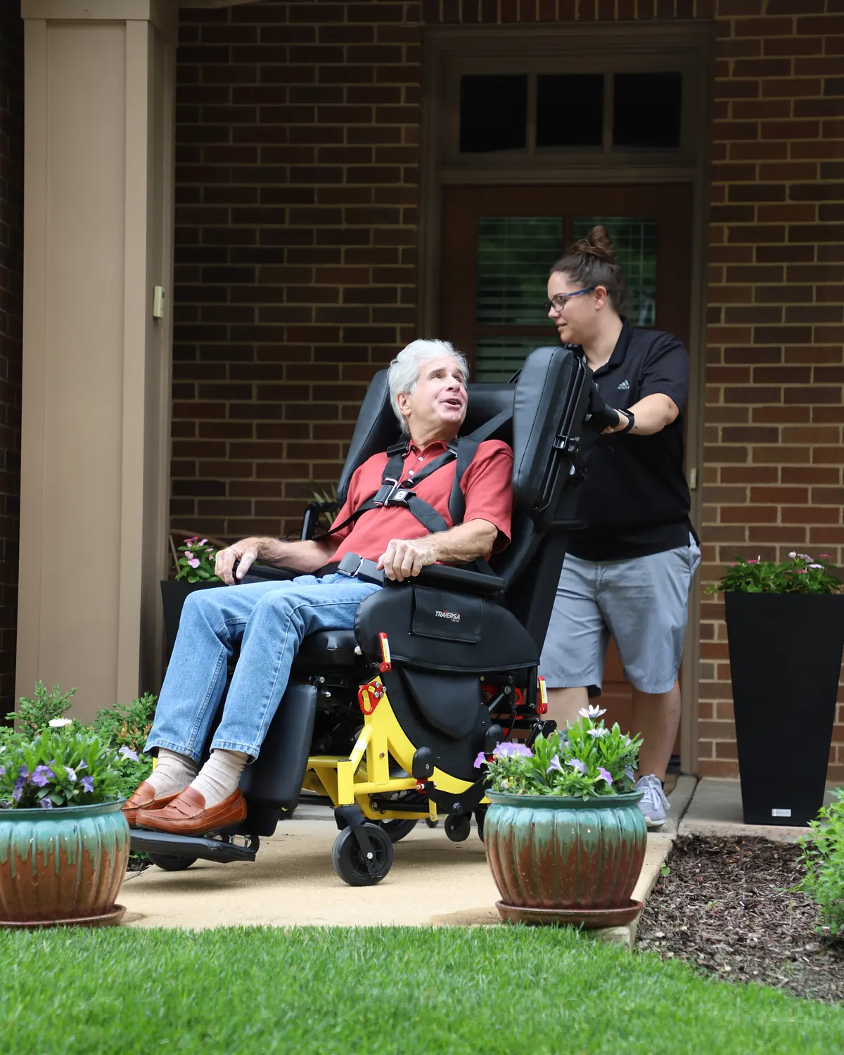 An older man sits tilted back in the Broda Traversa, the reclining transport chair Carolina Premier Mobility uses, looking up at the attendant behind him.