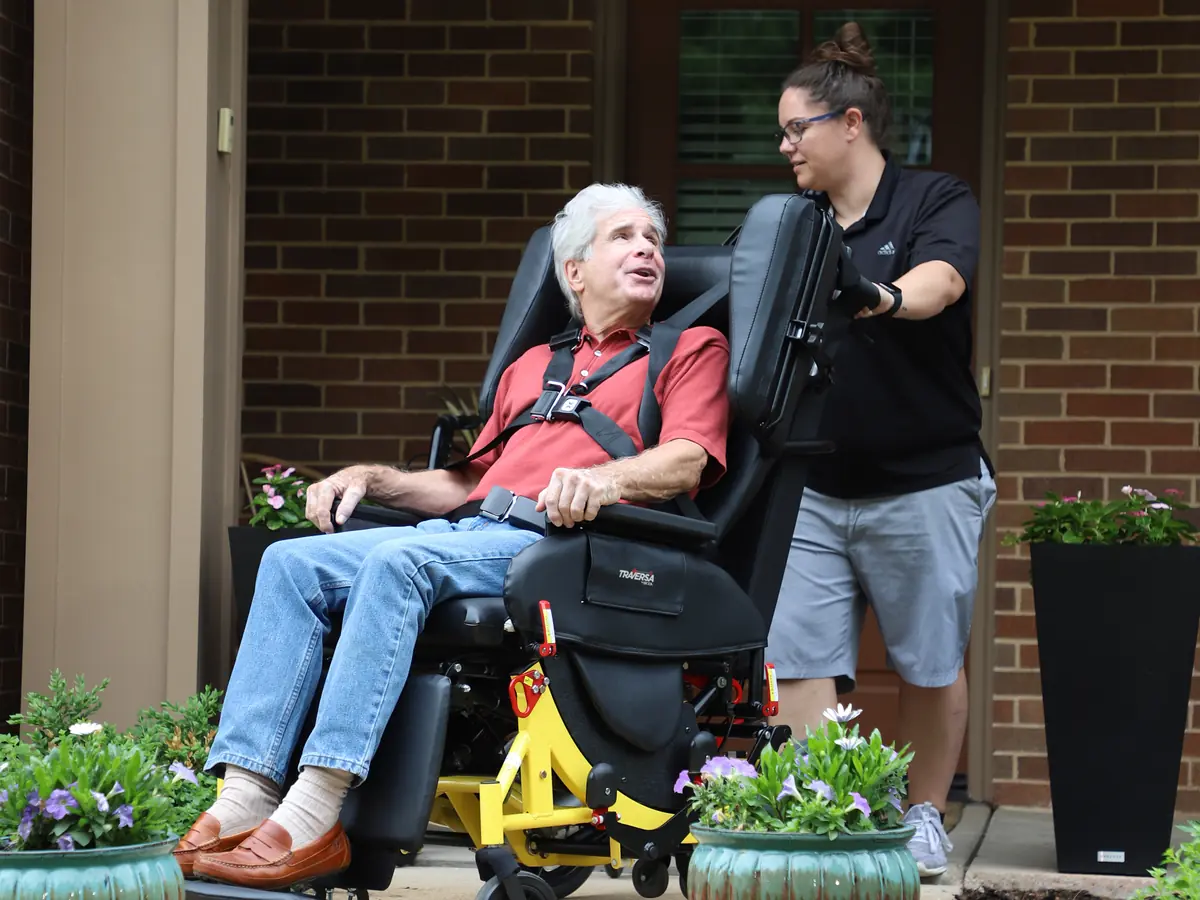 A rider sits buckled into the Broda Traversa reclining transport chair on a front walk, with an attendant at the handles.
